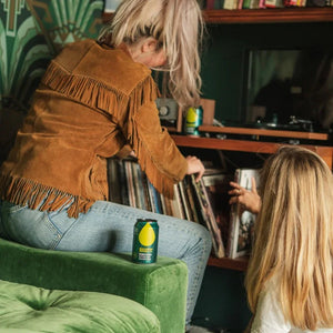 Two people are sitting on a green couch with a record player and records in the background, drinking Big Drop Paradsio Alcohol-Free-Citra-IPA