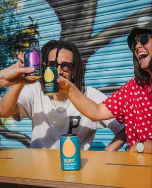 Two people holding cans of Big Drop Alcohol-Free Beer against a colorful mural.