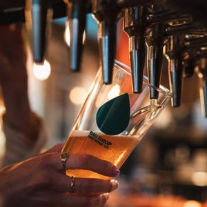 Person holding a glass of Big Drop alcohol-free beer being poured from a tap, with 'Big Drop Brewing Co.' branding.